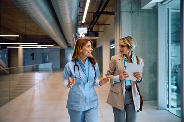 Happy nurse and businesswoman talking while walking through hospital hallway.