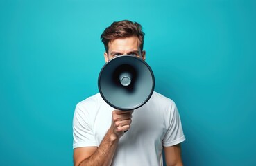Half portrait of young caucasian man shouting through megaphone on blue background. Guy hides face behind bullhorn, announcing important message. Concept of communication. Focus on eyes.