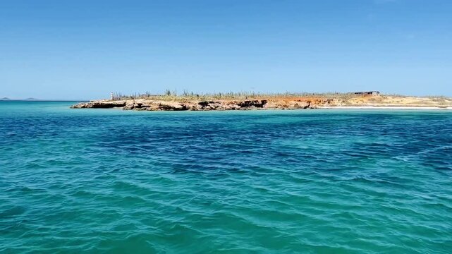 Crystal turquoise waters ripple under the sun as a catamaran glides along the rocky shores between Cubagua and Margarita Island, Venezuela. A tropical paradise with blue skies and vibrant seascapes 4K