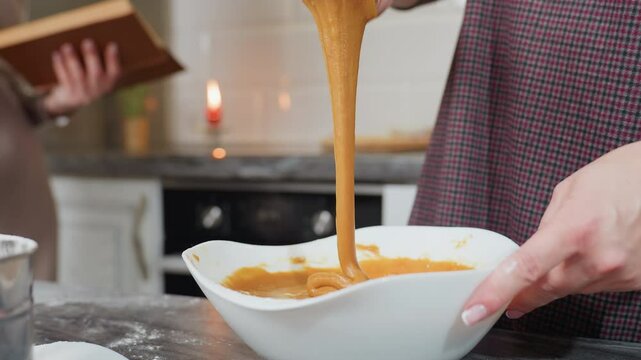 Close-up of woman holding whisk with thick caramel-colored pap stretching as it drips back into white bowl, kitchen background with blurred cookbook, candle, and flour-dusted countertop