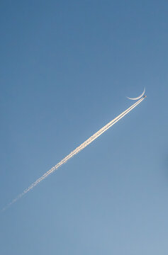 Airplane with contrail flying past crescent moon at dusk