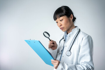 Medicine, healthcare, and people concept – female doctor with magnifying glass examining clipboard as if investigating, standing in front of an isolated background, focused and attentive.