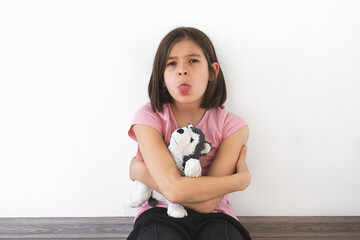 child girl 7-8 years in pink t-shirt sits against white wall, hugging plush toy and sticking out...