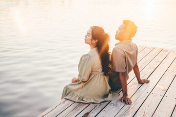 A young couple sits back to back on a wooden pier, gazing at the tranquil waters of the lake as the...