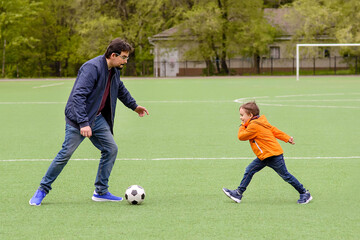 Obraz premium Father and toddler son having fun on green football field: dad and child playing football outdoors, kid attacking trying to kick soccer ball. Active happy family and healthy parenting concept