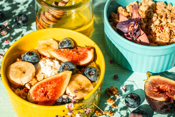 Granola with fresh banana, figs and blueberries on a light table with honey and flowers in a yellow and blue bowl. The concept of a delicious and healthy breakfast, a good start to the day