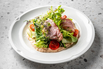 A fresh salad with mixed greens, cherry tomatoes, and slices of tender meat, elegantly presented on a white plate against a textured background.