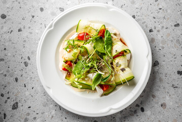 A fresh, colorful salad featuring sliced zucchini, cherry tomatoes, and mixed greens, arranged artfully on a white plate against a textured gray background.
