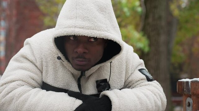 Rap artist in white hoodie and gloves leans on iron bar outdoors, nodding his head in thought, background features snowy ground, trees, and distant buildings