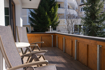 A wooden balcony with two reclining chairs and a table, overlooking modern buildings and tall evergreen trees in Ischgl, Austria's alpine setting.