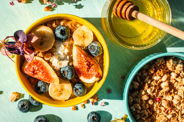 Healthy breakfast, figs, bananas and blueberries in yellow and blue bowls with yogurt and granola on a light table. View from above.