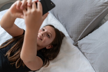 Fototapeta premium A young girl is lying on a cozy bed, holding a smartphone, and smiling while taking a selfie or making a video call. She wears a black t-shirt, surrounded by soft gray pillows in a relaxed setting.