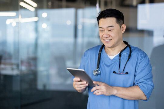 Smiling young Asian male doctor, assistant and nurse standing in the hospital lobby and using a tablet in his hands
