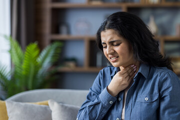 Close-up photo of a sick young Muslim woman sitting on the couch at home and holding her hand on her neck, feeling a severe sore throat