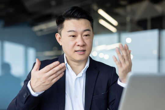 Asian young male businessman sitting in the office at the table and talking on a video call on a laptop, gesturing with his hands