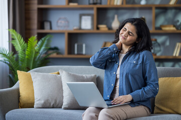 Tired young Indian woman sitting on sofa at home, massaging neck with hand, suffering from severe...