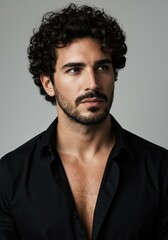 Obraz premium A handsome man with curly hair and a beard poses in a studio wearing an elegant black shirt.
