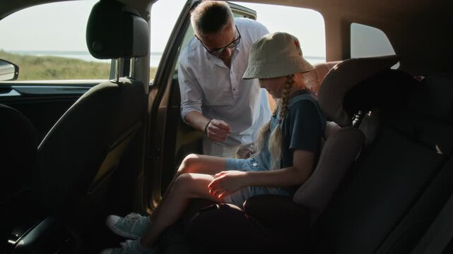 Young girl sitting in child safety seat in car, her father fastening her seatbelt before leaving lakeshore on summer day