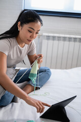 Naklejka premium Young woman sitting on a bed, knitting with soft pastel-colored yarn while using a tablet for guidance. Cozy and relaxing home activity focused on creativity and learning.