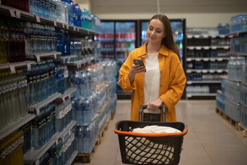 A woman walks through the supermarket, pushing a shopping cart. She checks her phone for a grocery...