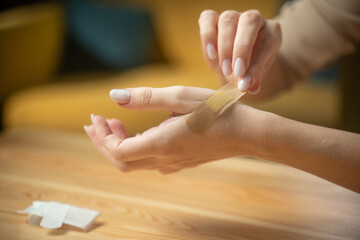 A young woman carefully places a Band-Aid on a small cut on her arm. She sits at a wooden table, demonstrating proper first-aid technique and self-care practices.