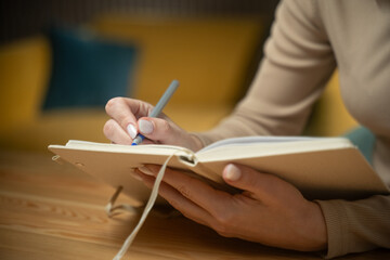 A person writes thoughtfully in a journal while seated in a warm, inviting space. The act of...