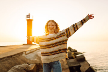 Young woman enjoying a breezy day at the beach near a vibrant red lighthouse at sunset. Travel,...