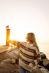 Young woman enjoying a breezy day at the beach near a vibrant red lighthouse at sunset. Travel,...