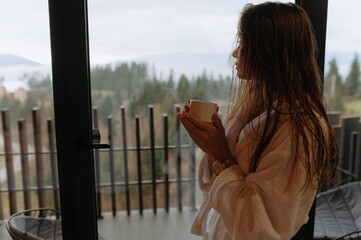 A woman in a robe enjoys a warm drink while gazing out at a beautiful mountain landscape on a serene morning.