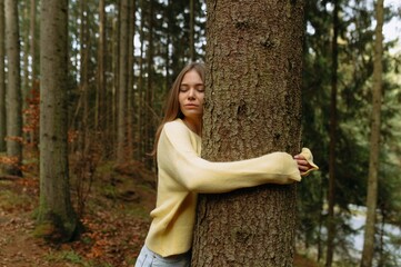 A young woman in a yellow sweater peacefully embraces a tree in a lush green forest, showcasing her...