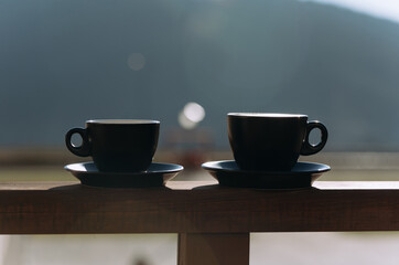 Two black coffee cups sit on a wooden balcony railing, with a beautiful blurred mountain view in the background, inviting a serene morning moment.
