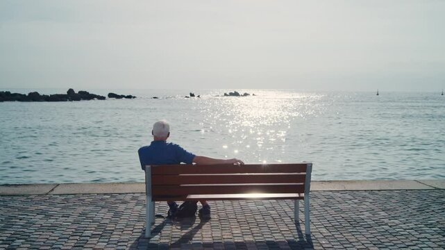 Old man sitting on a bench overlooking sea or ocean enjoying sunny day outside. Warm sun as senior male sits peacefully in a private quiet moment taking a break to look at view in vacation in Cyprus