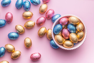 Chocolate easter eggs wrapped in multi colored foil in bowl on pink background. Top view.