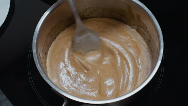 Close-up of cook stirring smooth pap on electric stove, creating a creamy texture with a partial view of a white plate, the thick porridge swirls as it is carefully prepared