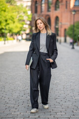 Thoughtful businesswoman with laptop walking through city streets, looking at camera. Freelancing,...