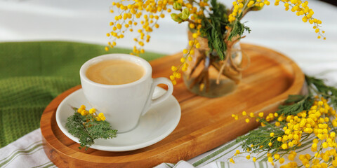 A white coffee cup on a wooden tray, decorated with fresh mimosa flowers, bringing a warm and festive spring morning feel.