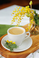 A cozy spring morning scene with a cup of coffee on a wooden tray, decorated with fresh mimosa flowers, creating a warm and festive vibe.