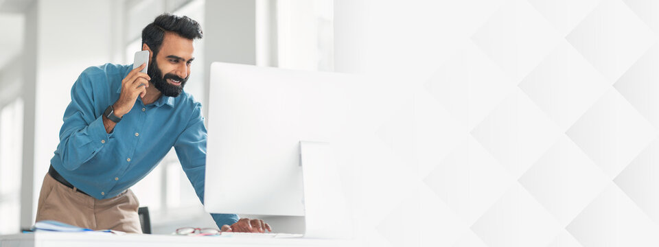 Cheerful bearded indian man in blue shirt multitasking, talking on phone and looking at computer monitor, working in light-filled contemporary office interior
