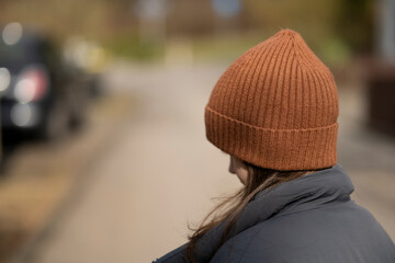 Fototapeta premium A girl with her back to the camera in a brown hat and jacket on the street on the path. Concept of children's loneliness and emotions