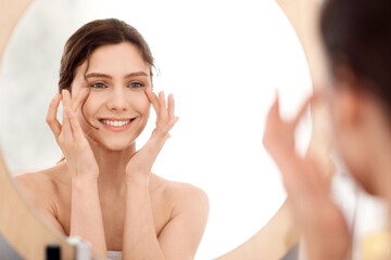 Eye care routine. Young woman massaging eye zone, looking at mirror, home interior