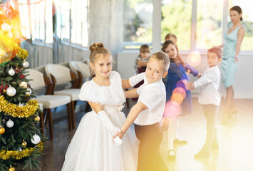 Couple of smiling tweens, boy and girl in festive clothing performing graceful waltz with group classmates under guidance of female teacher during Christmas event at school..