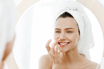Young woman looking at mirror and touching her nose, applying facial products after shower