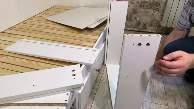 The man is assembling pull-out shelves for the sofa bed from wooden panels. He installs furniture cam locks.