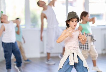 Energetic preteen girl practicing hip hop choreography in well-lit dance studio alongside peers and...