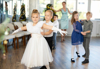 Smiling junior schoolchildren in party dresses learning waltz with pedagogue in hall embellished...