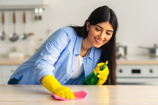 A young woman in yellow gloves smiles while cleaning the kitchen countertops. She uses a spray bottle and cloth, enjoying her housekeeping routine in a cozy home environment.