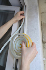 A man insulates windows and balcony doors with foam adhesive tape. Budget renovation in the apartment. Preparation for the cold snap. Heating problem in apartment buildings