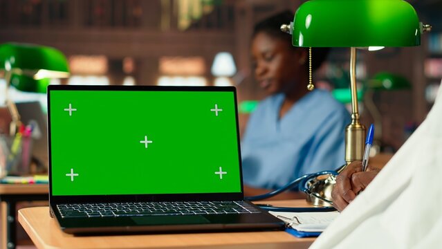 Med school student works on his thesis next to isolated mockup display in the library, doing medical research and reviewing specialty books. Future doctor studies with dedication. Camera B.