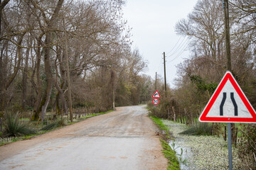 Road narrows sign on rural road with trees and cloudy sky. Dirt road with traffic signs and forest background. Countryside and pathways concept