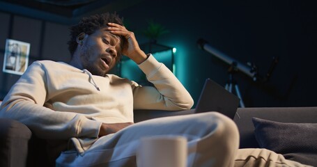Fototapeta premium Self employed man suffering from pain and high fever sitting on sofa, trying to work and finish his tasks. Black male feelancer suffering from illness and feeling sick, using laptop. Camera A.
